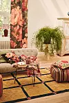 Ljust vardagsrum med soffa, blommiga gardiner, stora gröna växter, randig sittpuff och rutmönstrad matta.
