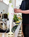 Vit stavmixer står på köksbänken med grönsaksskål och kastrull i bakgrunden.