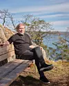 En man sitter på en träbänk på en klippa med hav och träd i bakgrunden.