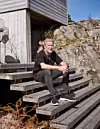 En man i svart sitter på en trätrappa utanför ett modernt hus på klipporna med havet i bakgrunden.