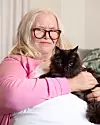 En kvinna med glasögon och rosa tröja håller en svart katt i knät i en soffa.