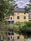Gult hus med svart tak speglas i stilla vatten omgiven av grönska.