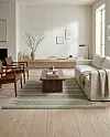 Ljust vardagsrum med beige soffa, lågt träbord, trästolar, matta och stor vas med grenar.