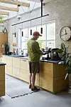 Person vid köksbänken i ett modernt kök med stora fönster, växter och pendellampor.