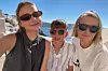 Tre personer med solglasögon står framför havet och vita byggnader en solig dag.