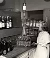 Svartvit bild av en kvinna vid en bardisk med flaskor, glas och en tappkran.