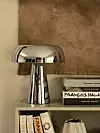 Kromad bordslampa på hylla framför böcker och brun väggbonad.