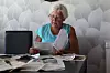 Kvinna sitter vid ett bord med tidningar och dokument i ett ljust rum med mönstrad tapet.