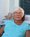 Äldre kvinna med glasögon sitter på en altan framför ett fönster.