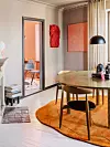 Rundt träbord, orange matta och svart lampa i en varm, modern matplats med öppen dörr i bakgrunden.