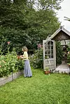 En person vid en blomsterbädd bredvid ett litet växthus i en grön trädgård.