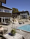 Modernt hus med trädeck, pool, solstolar och sittgrupper i en ljus trädgårdsmiljö.