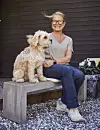 Kvinna sitter på en bänk med en ljus hund framför en mörk trävägg.