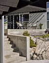 Modern terrass med stolar, bord och krukväxter intill ett hus med glasväggar.