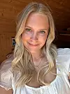 Närbild av en leende blond kvinna i vit blus framför en trävägg.