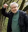 En äldre man med glasögon står utomhus bland träd och höjer ena armen.