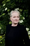 En kvinna i svart klädsel står framför en vägg av gröna blad och vita blommor.