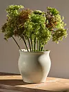 Stor bukett med gröna och rödlila blommor i en ljus keramisk vas på ett träbord.