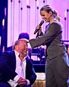 Kvinna med mikrofon står böjd mot sittande man på scen med lila ljus.
