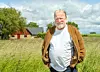 En man står i ett gräsfält framför ett hus, med sädesfält och molnig himmel bakom.