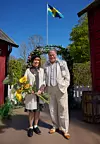 Två personer står framför en blomsterbåge på en gård med svensk flagga i bakgrunden.