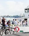 Flera personer går ombord på en färja vid kajen med stadssilhuett i fjärran.
