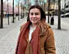 En kvinna står på en trottoar med träd och byggnader i bakgrunden.