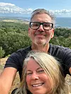Två leende vuxna tar en selfie framför ett kustlandskap med skog, hav och molnig himmel.