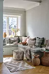 Ljus sittplats vid fönster med kuddar, plädar, puff, matta och flätade korgar.