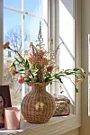 Blommor i en stor korgvas står i ett soligt fönster med ljus och en liten dekorativ ask bredvid.