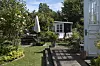 Solrig kolonihave med træterrasse, havemøbler, blomsterbede og lille hvidt havehus i baggrunden.