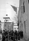 En grupp människor står samlad utanför en stor byggnad med en flagga som hänger från fasaden.