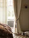 Ljust sovrum med öppet fönster, beige gardiner, en sängkant och en dekorationsvas på väggen.