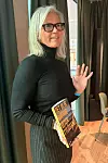 Kvinna med glasögon och grått hår håller upp en bok och vinkar i ett rum med gardiner och trägolv.