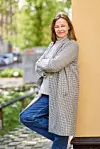 En kvinna står utomhus lutad mot en gul vägg med korsade armar och jeans.