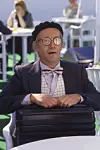 Man i kostym och basker sitter med en svart resväska vid ett bord utomhus.