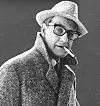 Svartvitt porträtt av en man med hatt, glasögon och cigarett mot neutral bakgrund.