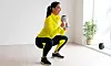 Kvinna i gul jacka gör knäböj med kettlebell i ett ljust rum.