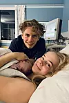 Nyfødt baby ligger hos mor i sykehusseng mens en annen voksen smiler mot kameraet.