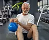 Äldre man i vit t-shirt håller en blå kettlebell i ett gym.
