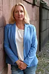 En kvinna står lutad mot en stenmur på en smal gata, klädd i blå blazer och jeans.