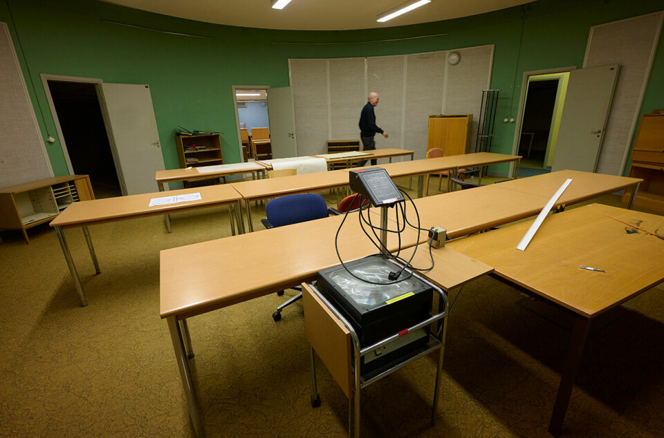 Tomt mötesrum med bord, stolar, projektor och gröna väggar i en bunkerliknande lokal.