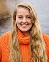 Leende kvinna med långt blont hår i orange stickad tröja mot oskarp bakgrund.