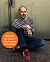 En man i randig tröja sitter på marken mot en vägg med röda skor och ett orange informationsmärke.