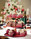 Tre våningar kakfat med cupcakes och pepparkakshus på ett dukat bord med julig bokeh-bakgrund.