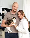 Ett par håller en katt i ett ljust rum med gardiner och en klösmöbel i bakgrunden.