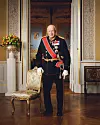 Kung Harald V i svart galauniform med ordnar, stående i ett rikt dekorerat rum bredvid en stol.