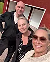 Tre personer står utomhus framför en röd byggnad och poserar för en selfie.