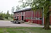 Röd gårdsbyggnad vid grusplan med släp och träd i förgrunden.