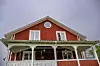 Röd träbyggnad med vit veranda och rund gavelprydnad under en grå himmel.